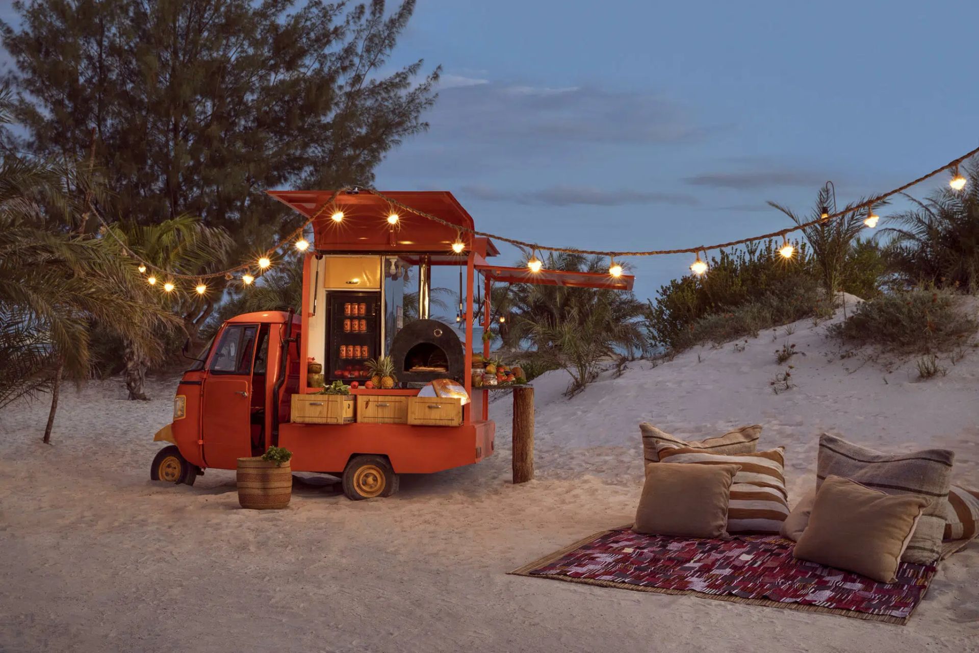 An orange food cart parked on a sandy beach at dusk, illuminated by string lights with cushions on a rug in the foreground.