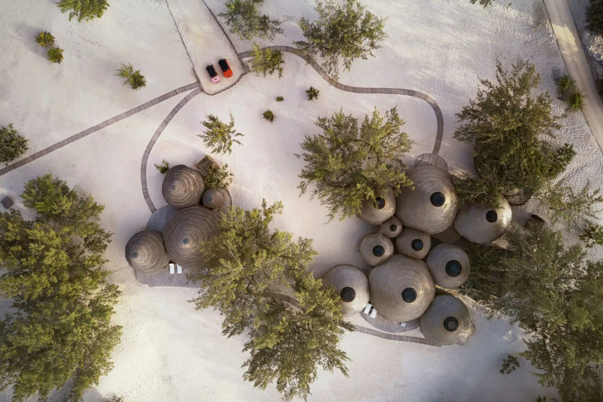An aerial view of a cluster of domed, textured structures nestled among trees on a light-colored, sandy ground.