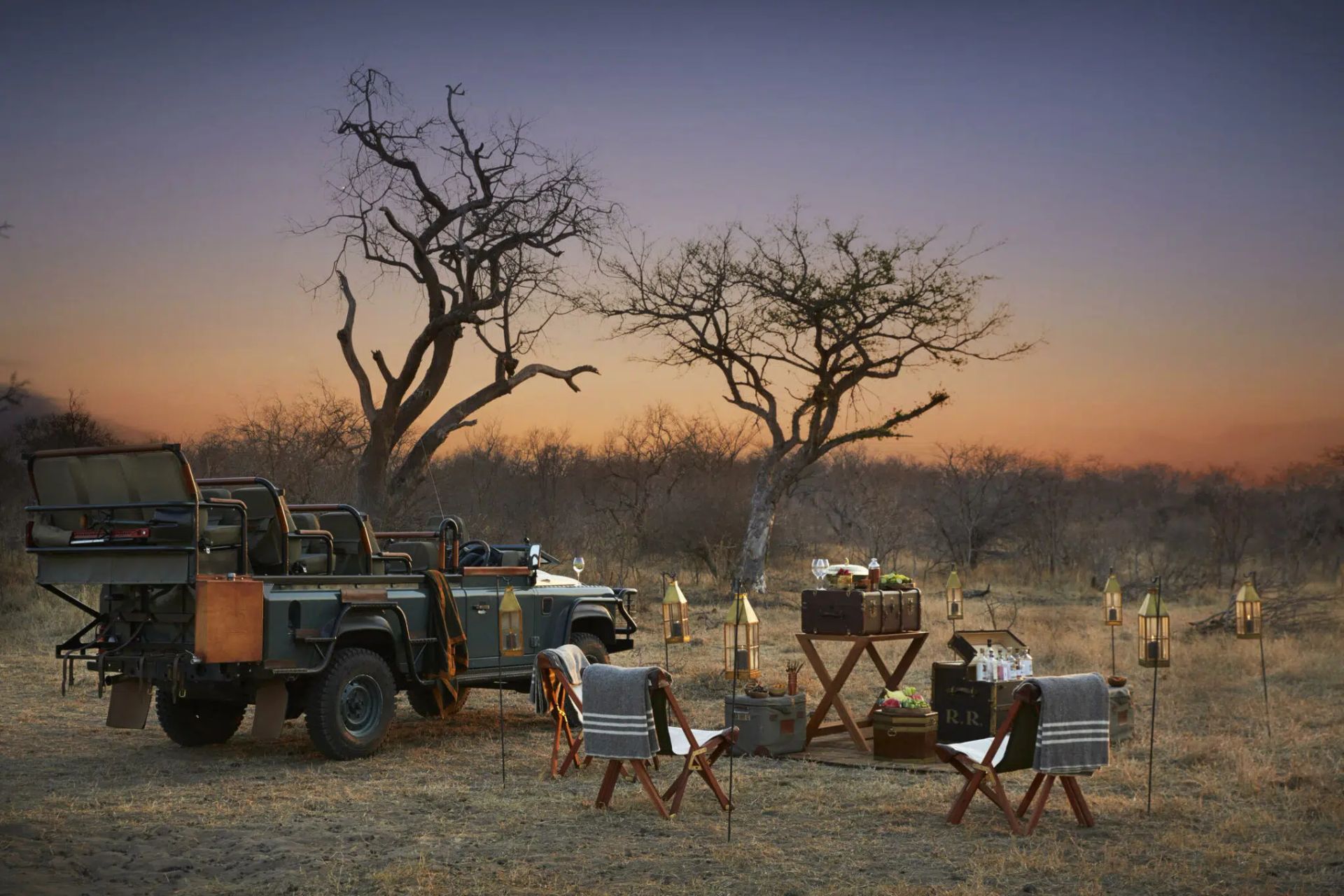 Safari camp at sunset with vehicles, tents, and two bare trees in a dry field