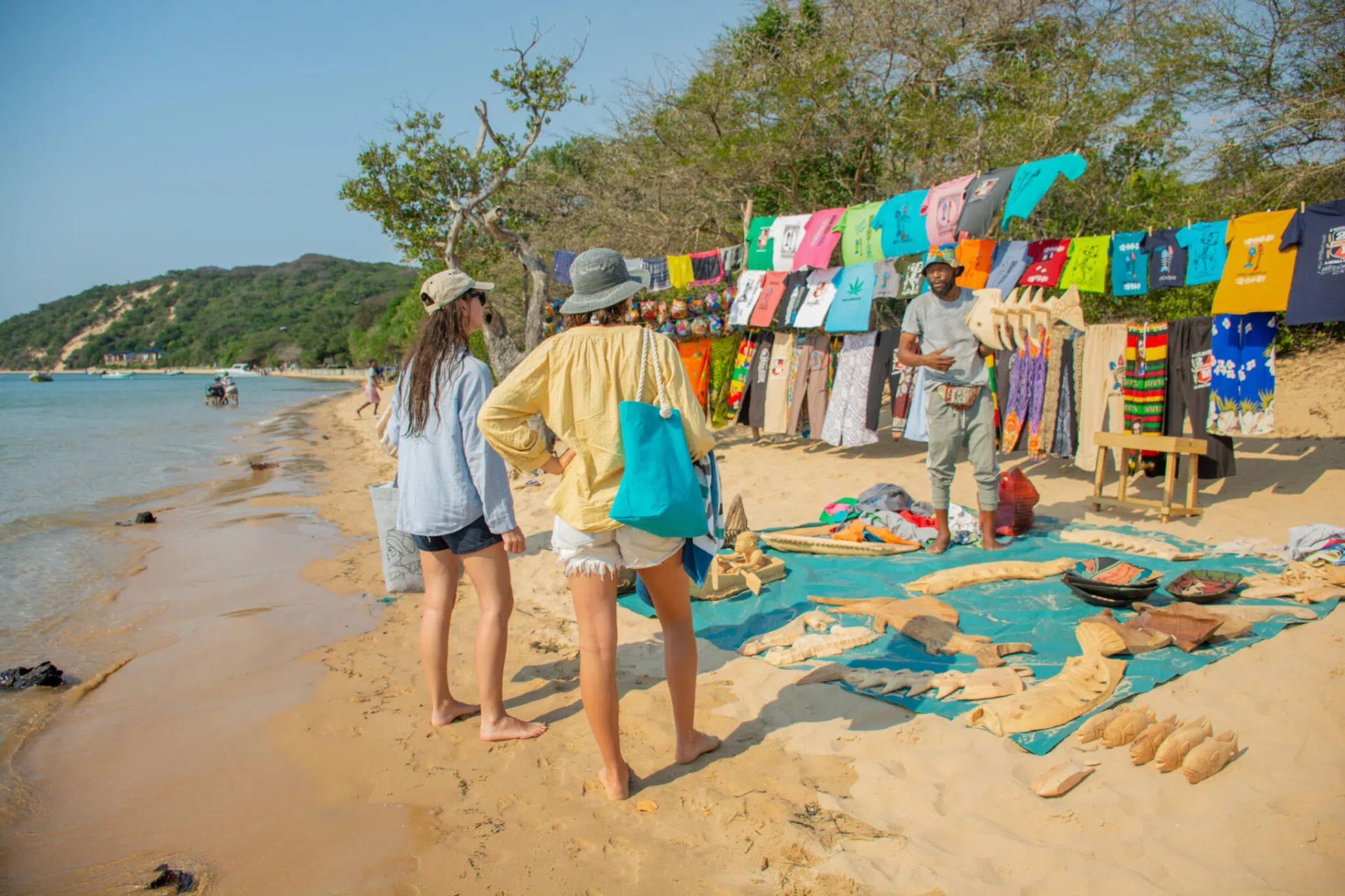 People browsing colorful beach clothing stalls beside a sandy shore, with towels and trees nearby.