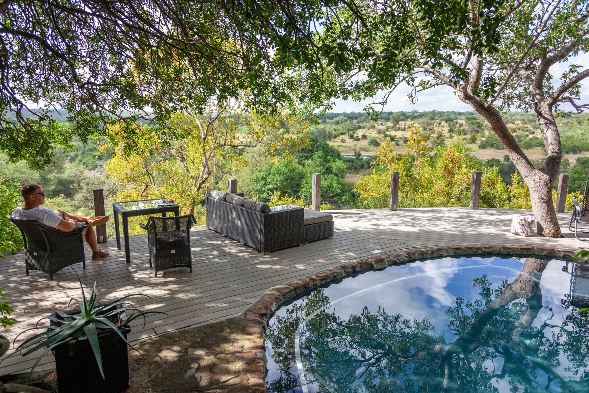 Patio with outdoor seating beside a pool, shaded by trees overlooking a green valley