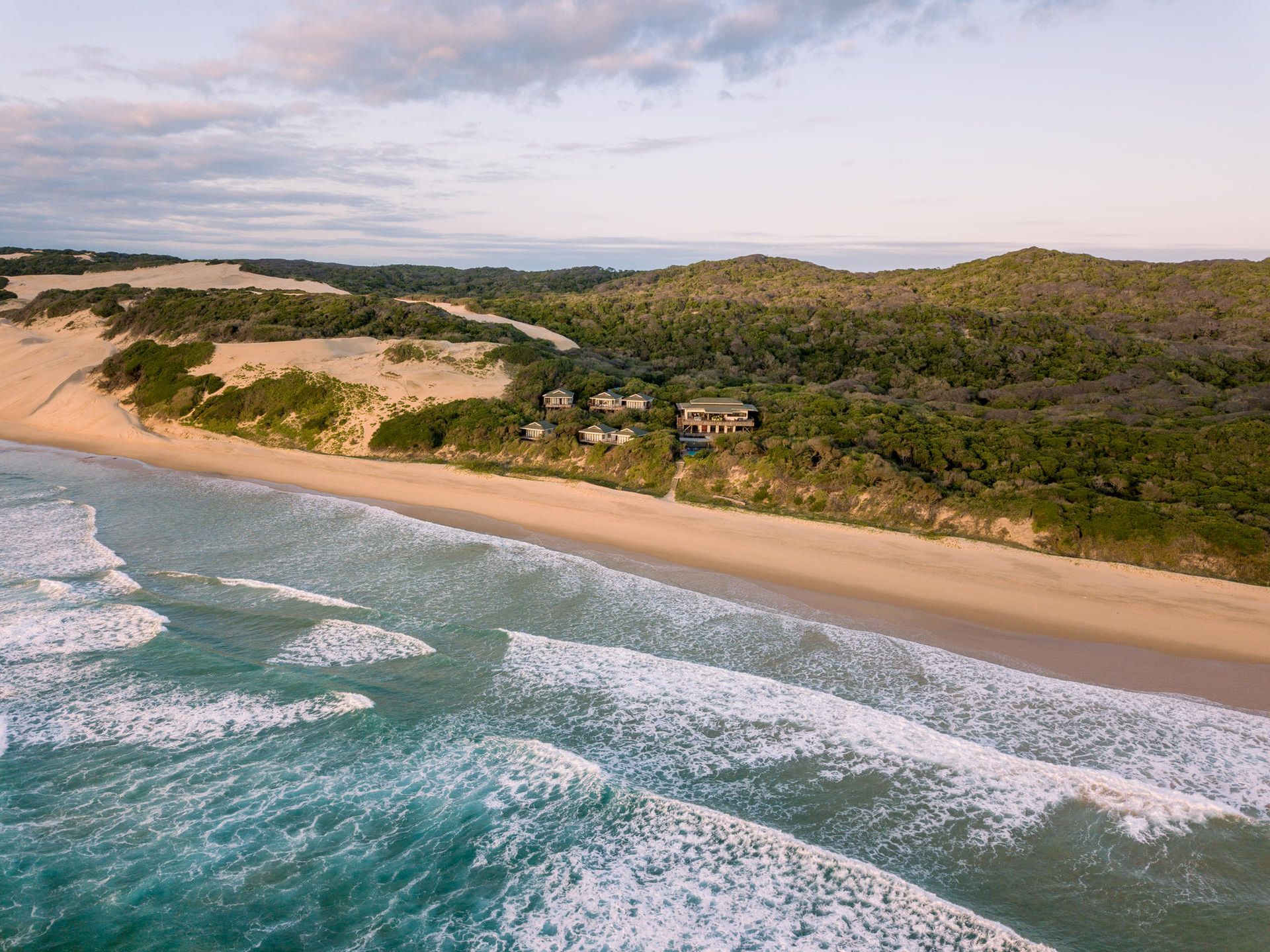 Aerial view of a sandy beach with turquoise waves, green dunes, and a rugged coastline.