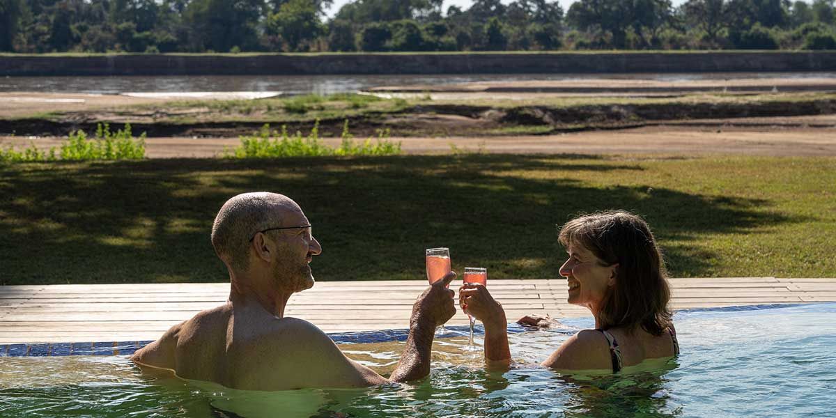 Two people in a pool clink drinks at a resort with a river and trees in the background