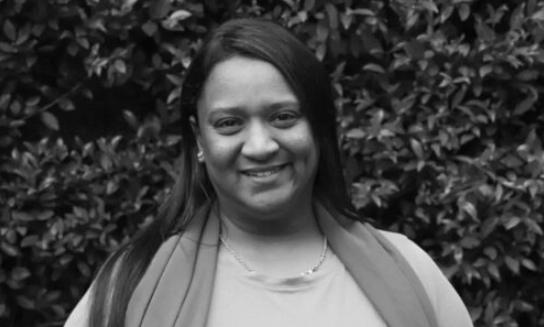 Smiling person in a light scarf standing before leafy greenery.