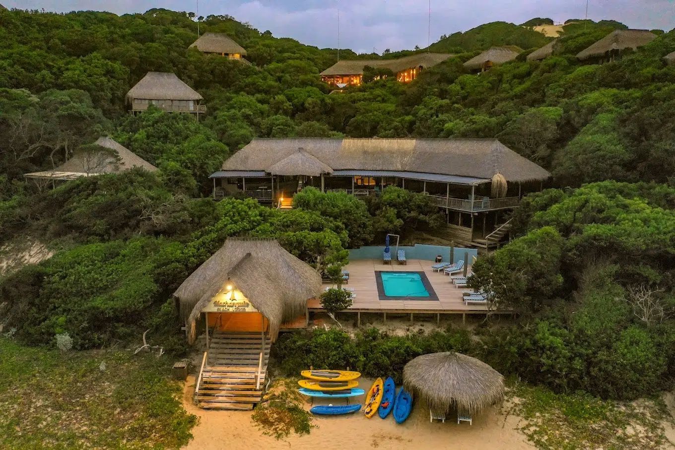 Tropical resort with thatched-roof huts, a central pool, and lush green hillside vegetation