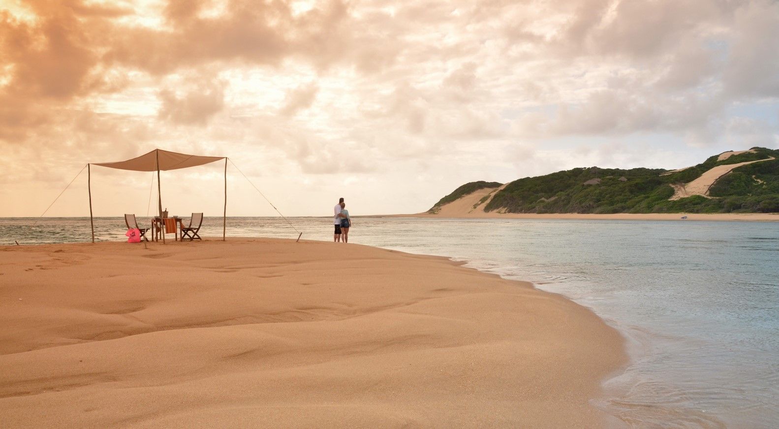 Sandy beach with a canopy, a few people, calm water, and a green headland under a cloudy sunset sky