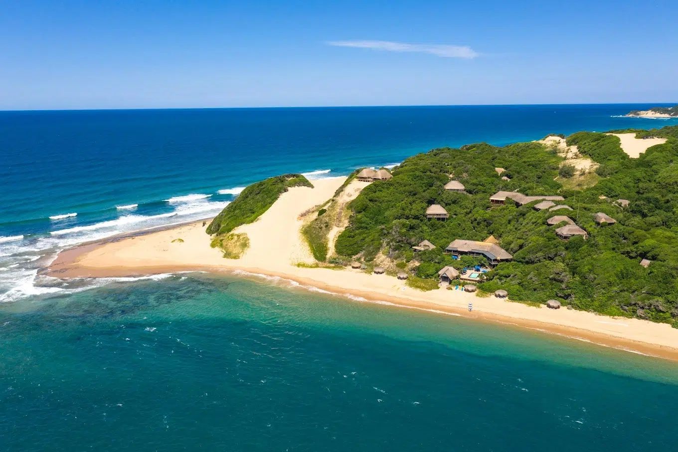 Aerial view of a sandy tropical beach with turquoise water, green vegetation, and small huts along the shore