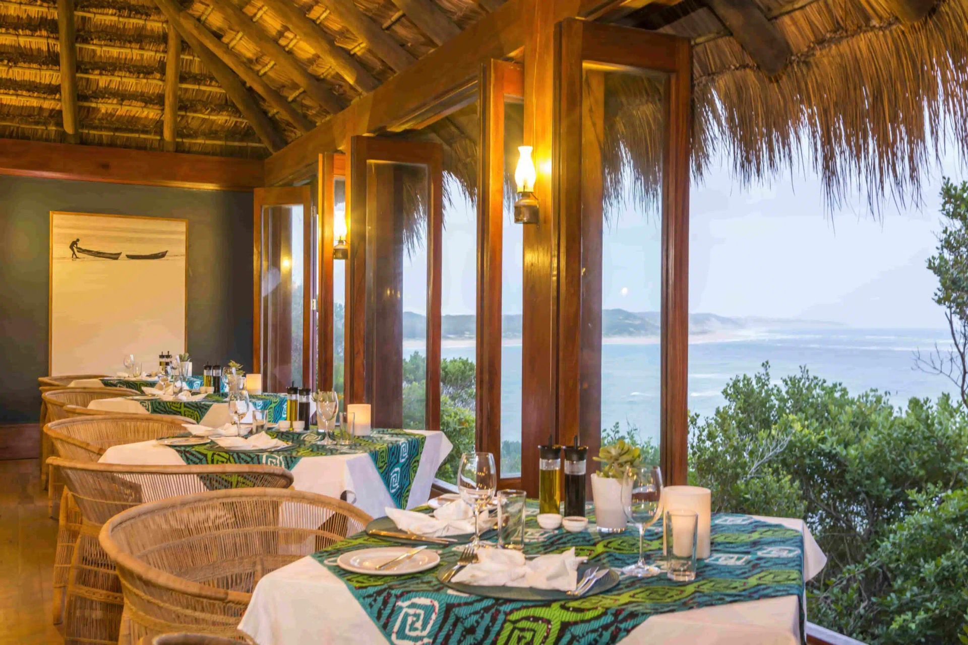 Open-air seaside restaurant with set tables, wicker chairs, and ocean views at sunset