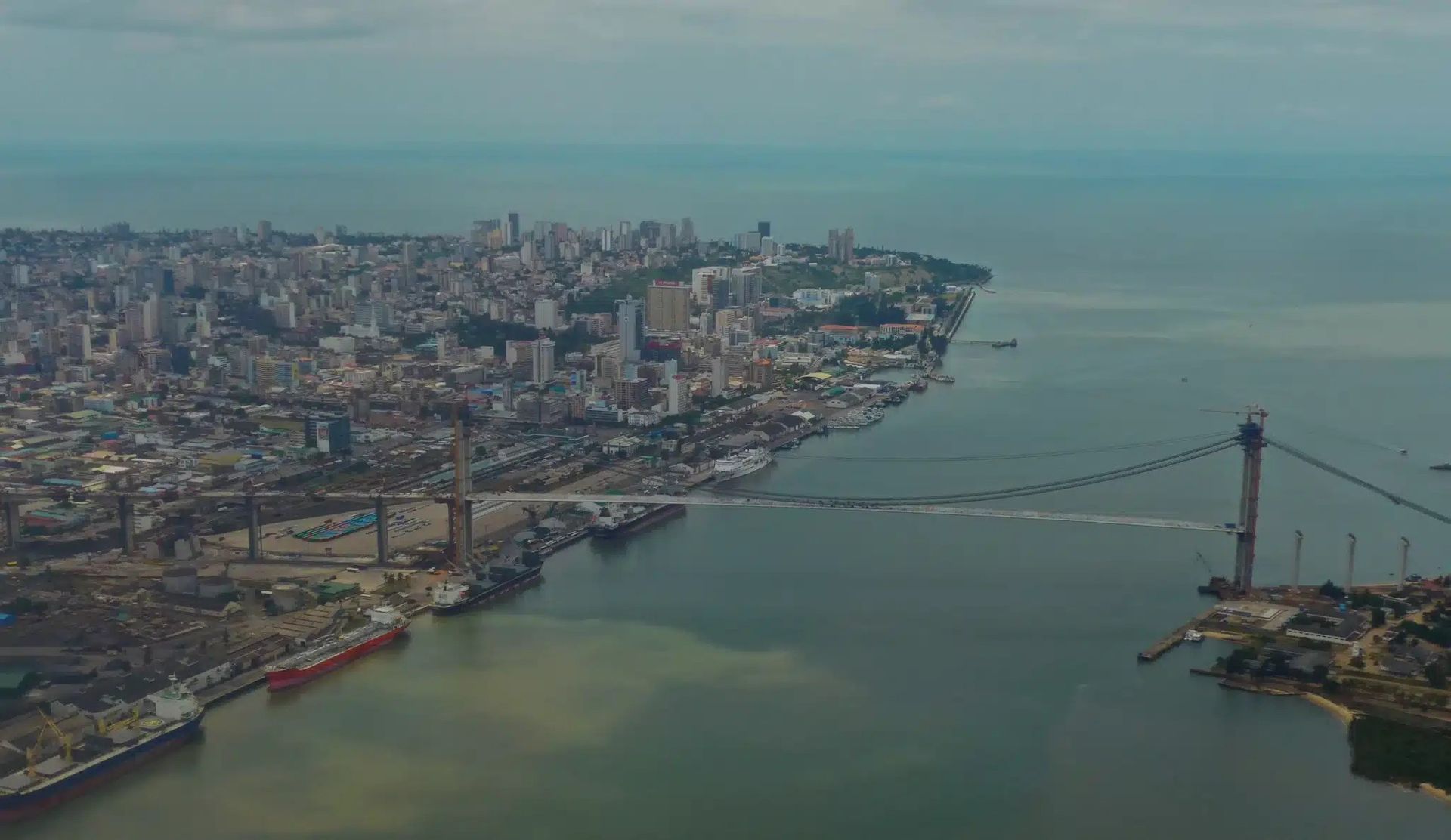 Aerial view of a dense city along a wide river, with a long suspension bridge crossing the water.