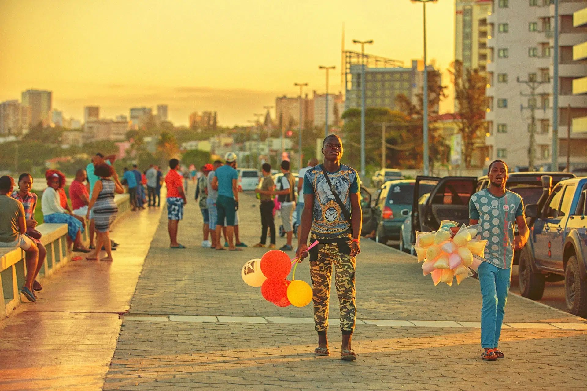 Two people walk along a sunset-lit waterfront promenade, one carrying balloons, with parked cars and pedestrians nearby.