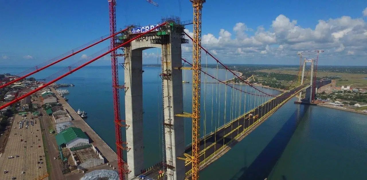 Aerial view of a large suspension bridge under construction over water with cranes and cables.