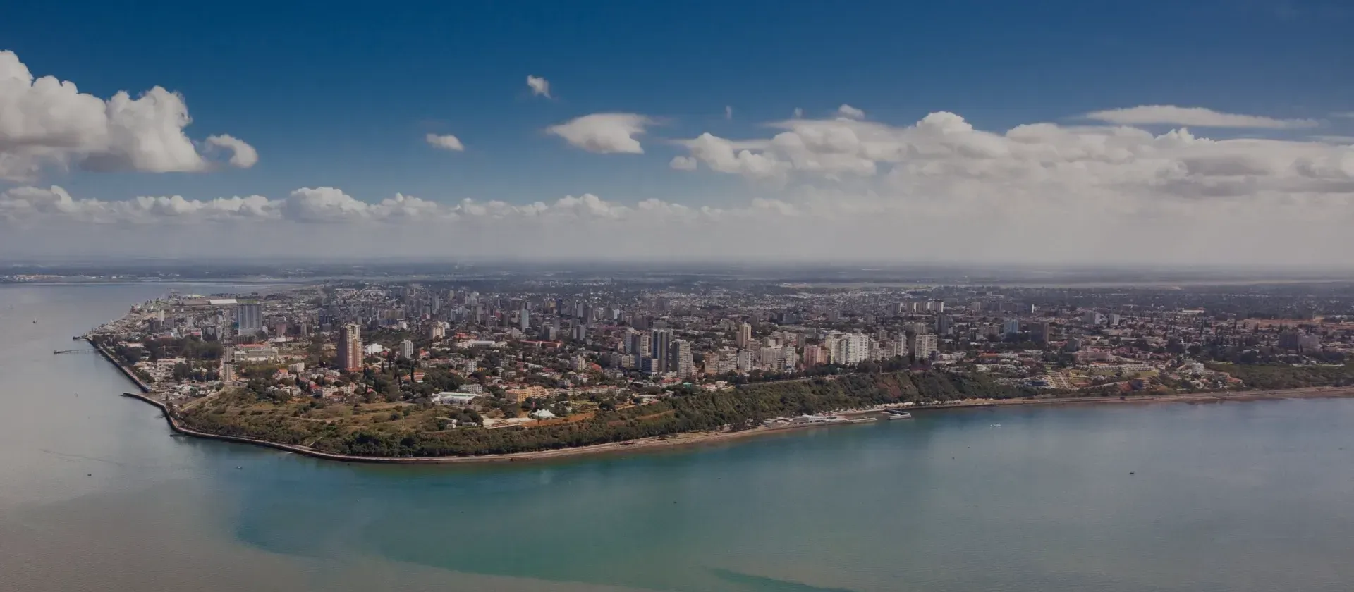 Aerial view of a coastal city skyline beside a wide river under a cloudy blue sky.