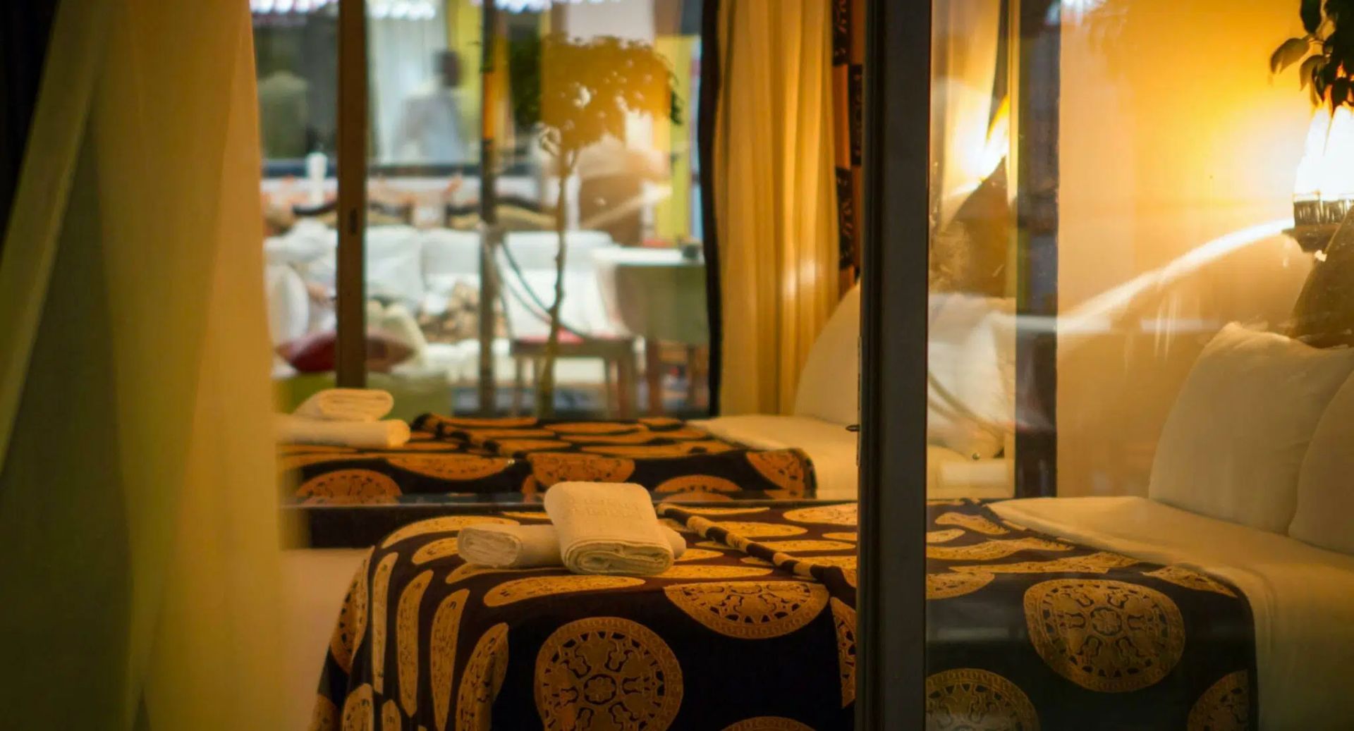 Warmly lit bedroom with a bed, patterned bedding, curtains, and a view through glass doors to a patio.