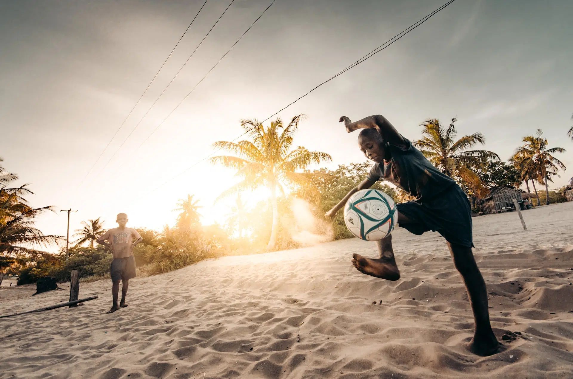 Person kicking a soccer ball on a sandy beach at sunset, with palm trees in the background