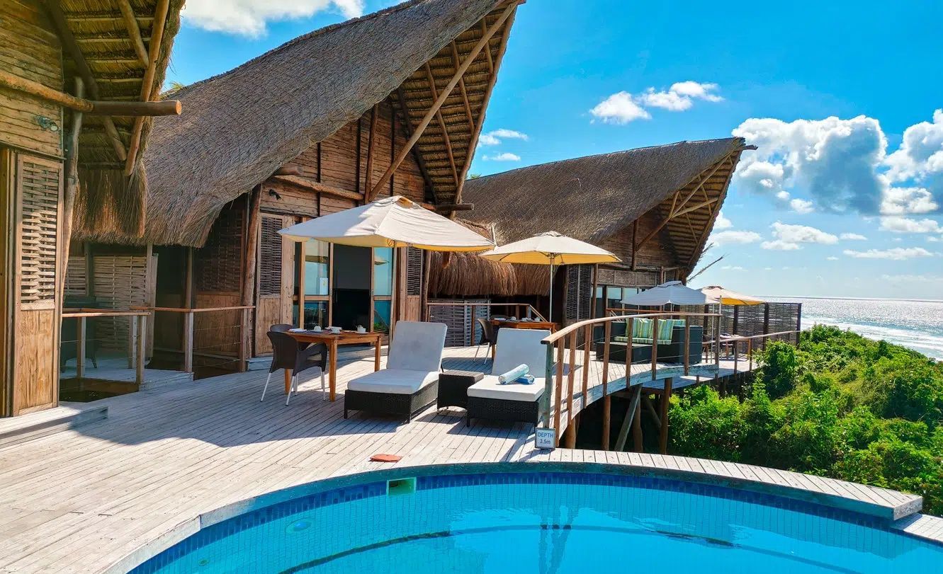 Luxury resort terrace with pool, lounge chairs, umbrellas, and thatched-roof villas overlooking lush greenery and sky