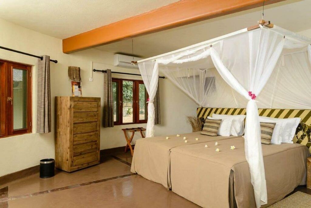 Bedroom with four-poster bed, beige bedding, wooden dresser, and windows in a bright room