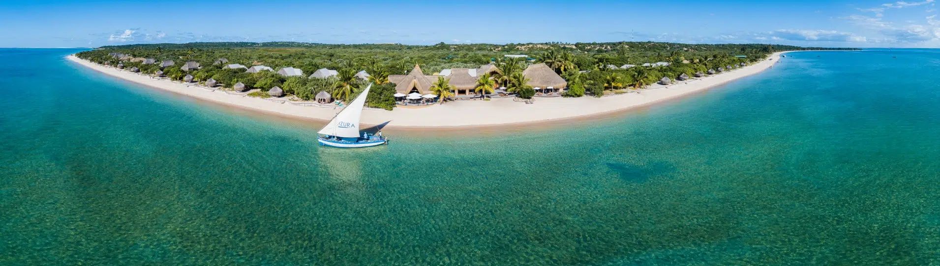 A wide aerial view of a sandy beach resort, tropical greenery, turquoise water, and a traditional dhow boat docked nearby.
