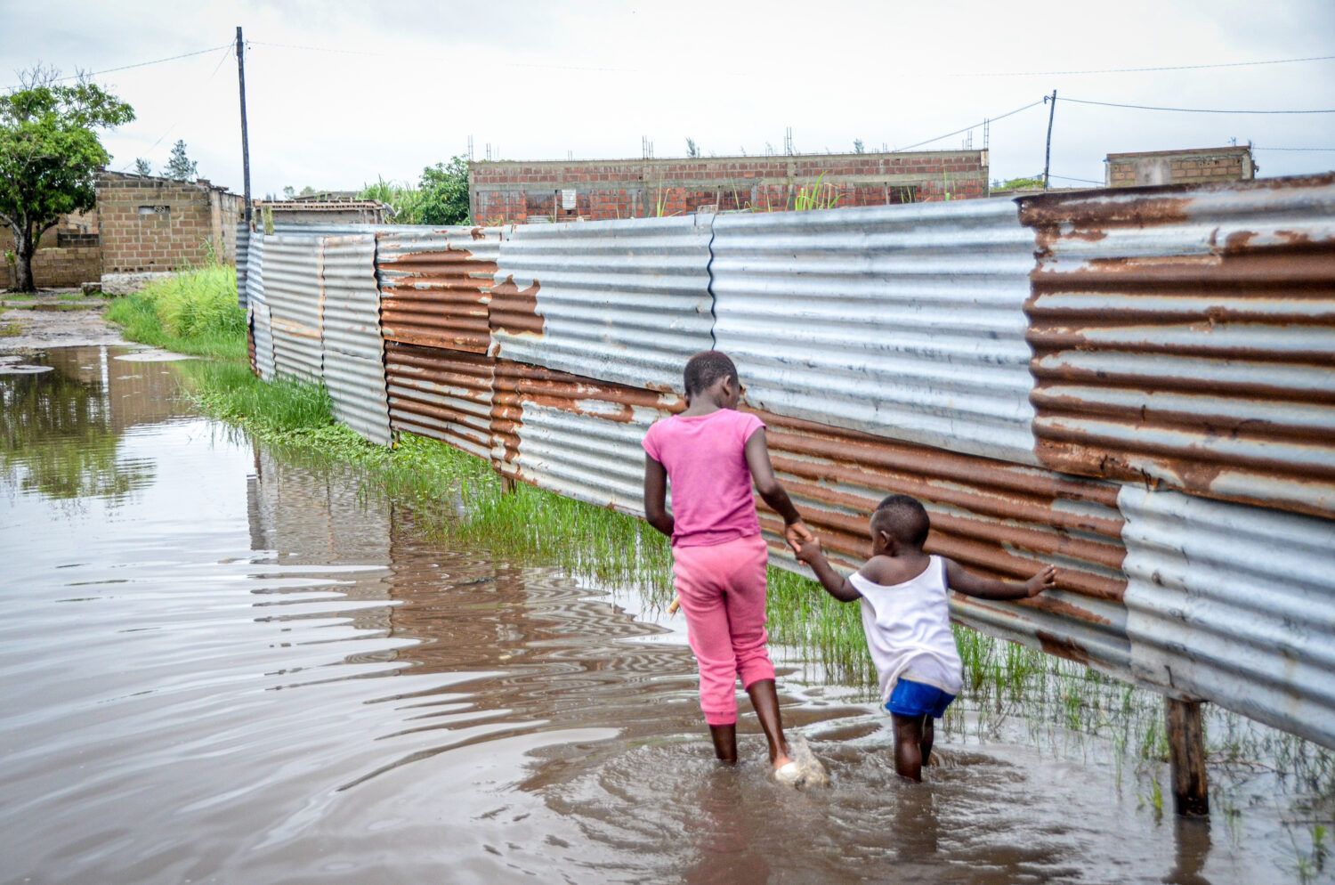 Two people wading through floodwater beside a corrugated metal fence.