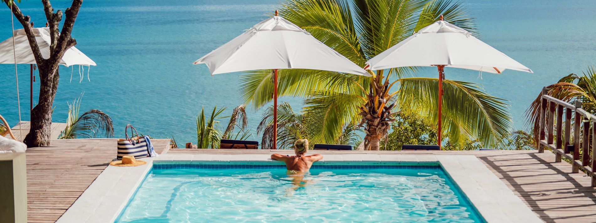 Tropical resort pool with sun umbrellas, palm trees, lounge chairs, and ocean view