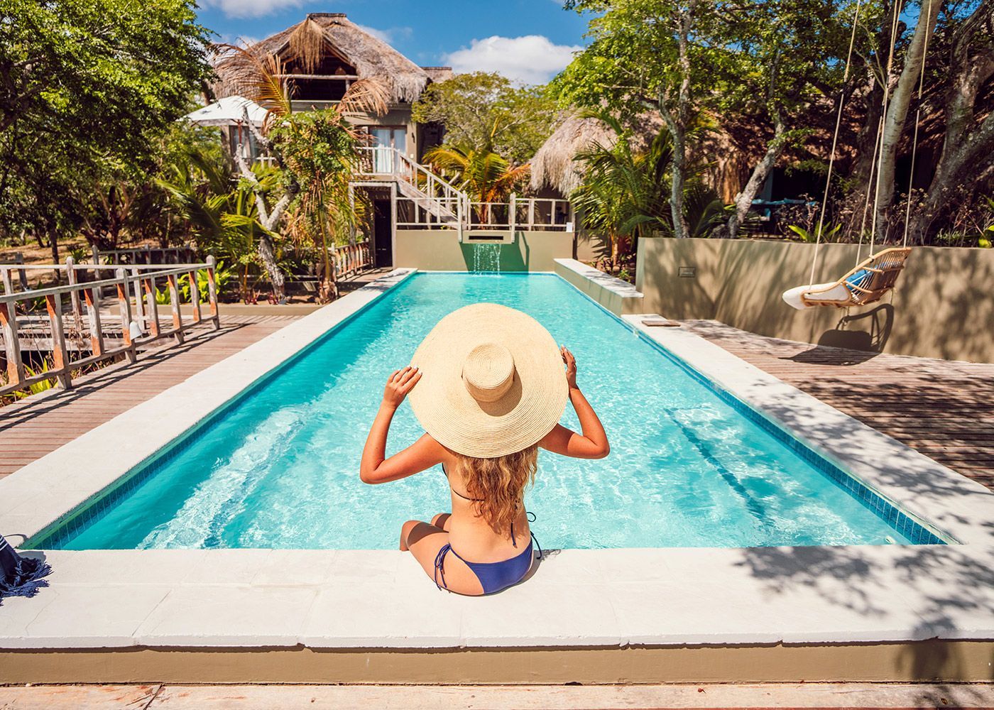 Woman in a straw hat sitting by a turquoise resort pool surrounded by tropical trees and thatched buildings