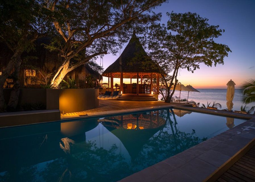 Tropical resort pool at sunset with a lit pavilion, palm trees, and ocean view