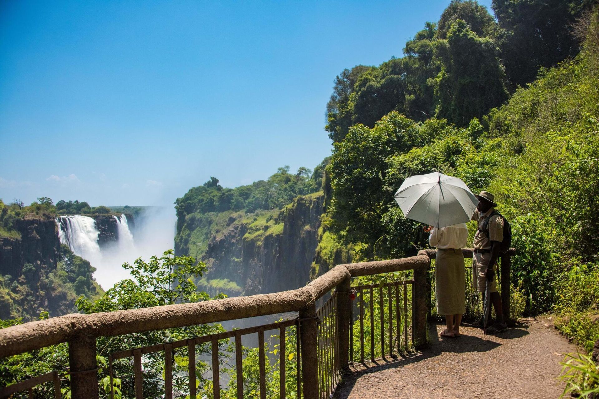 Viewpoint overlooking a waterfall gorge, with a person holding a white umbrella beside a wooden railing.