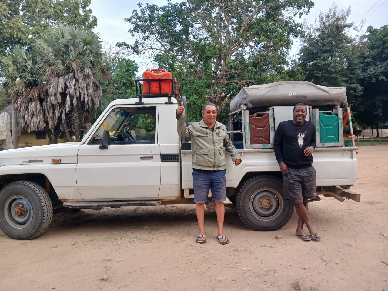 Two men standing beside a white pickup truck on a dirt road in a rural setting