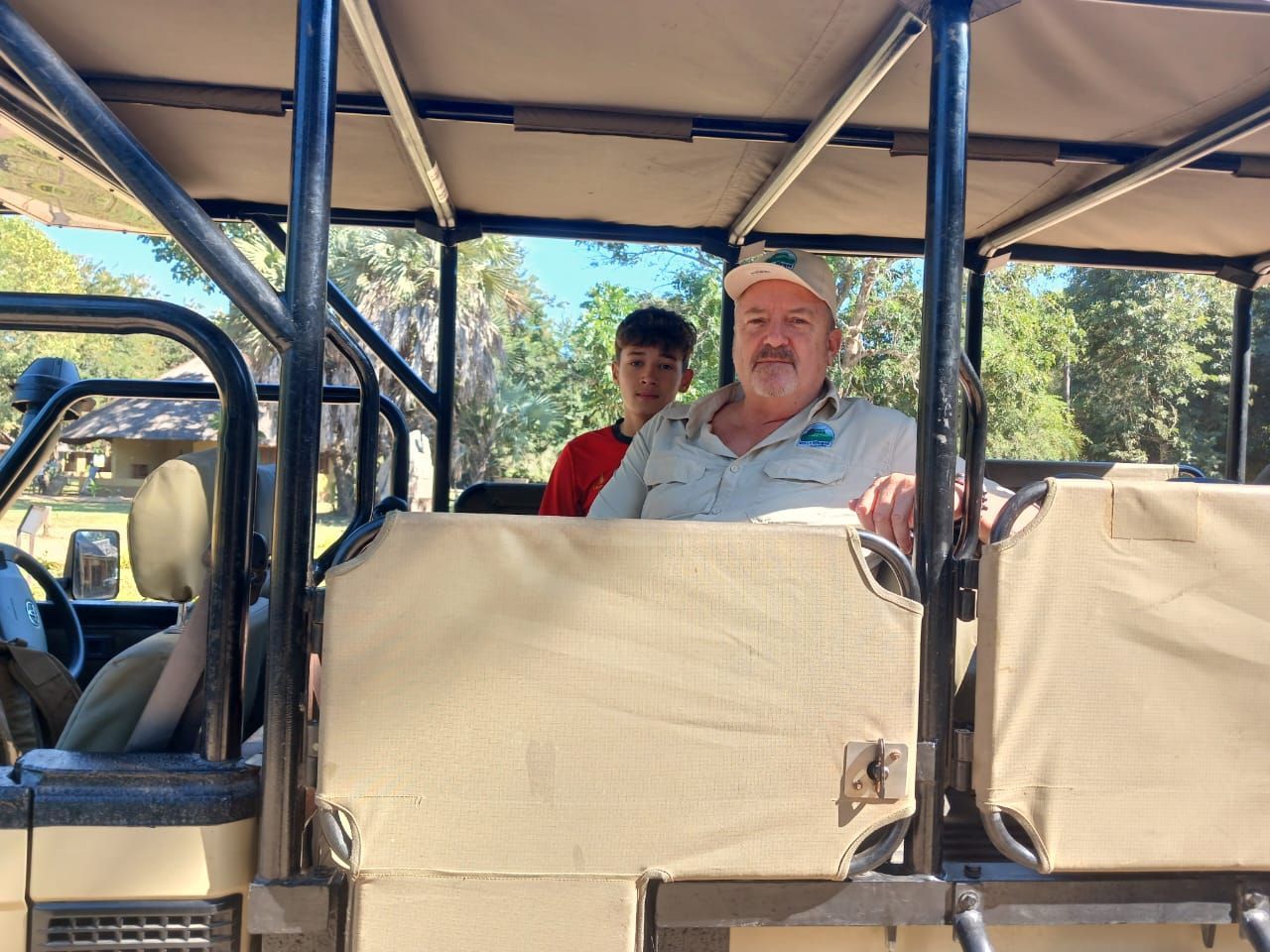 Two people seated in an open safari vehicle under a canopy, with trees in the background.