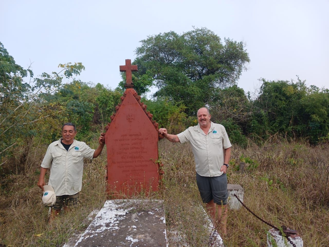 Two people stand beside a red roadside shrine with a cross, in a grassy rural area.
