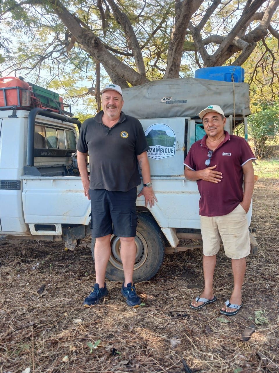 Two men standing by a white truck under trees, one smiling and one giving a thumbs-up.