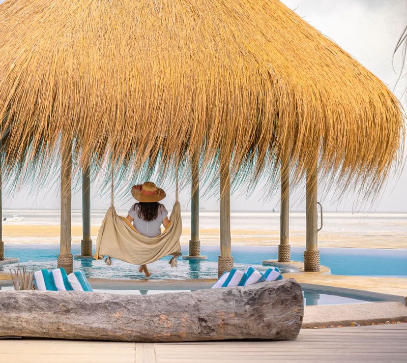 Thatched beach cabana with hanging hammock, poolside loungers, and ocean view at sunset