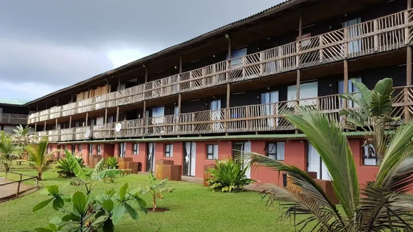 Two-story wooden lodge with balconies and red doors, seen across a green lawn under cloudy skies