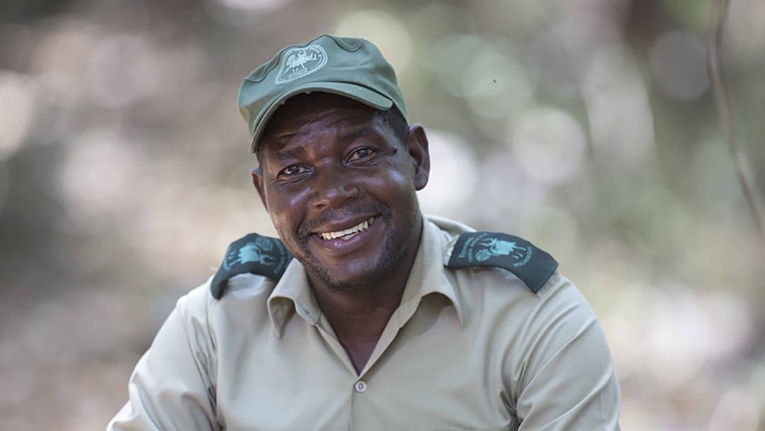Smiling park ranger in a khaki uniform and green cap, seated outdoors with blurred trees behind him