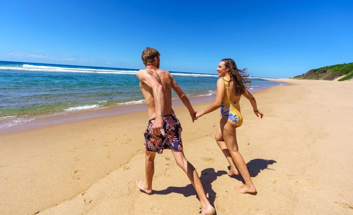 Two people holding hands while walking barefoot along a sunny beach beside the ocean