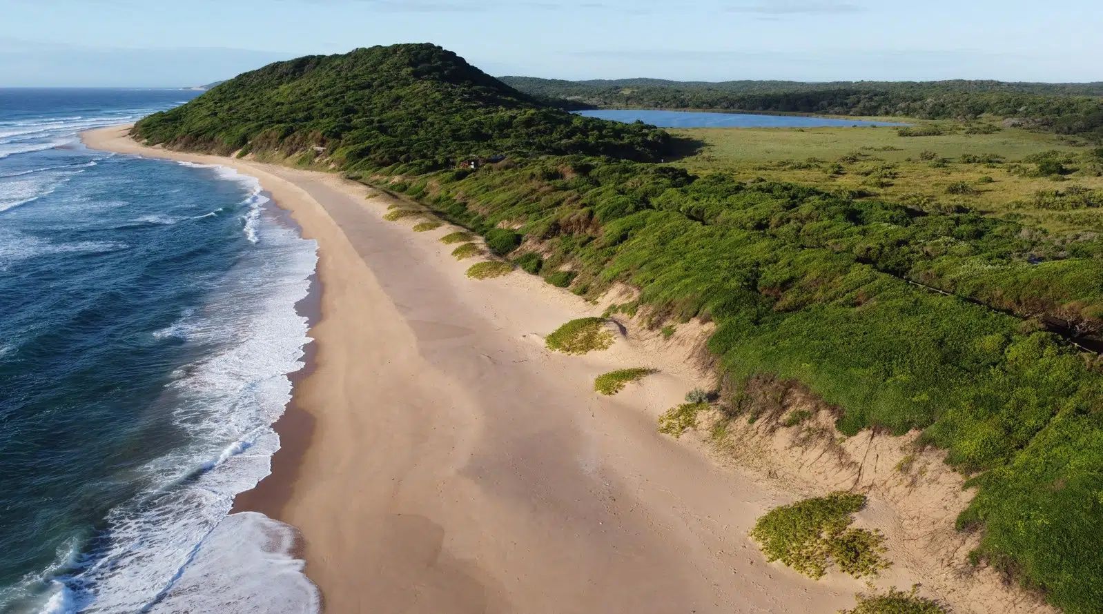 Aerial view of a sandy beach beside blue ocean waves and green coastal hills under a clear sky