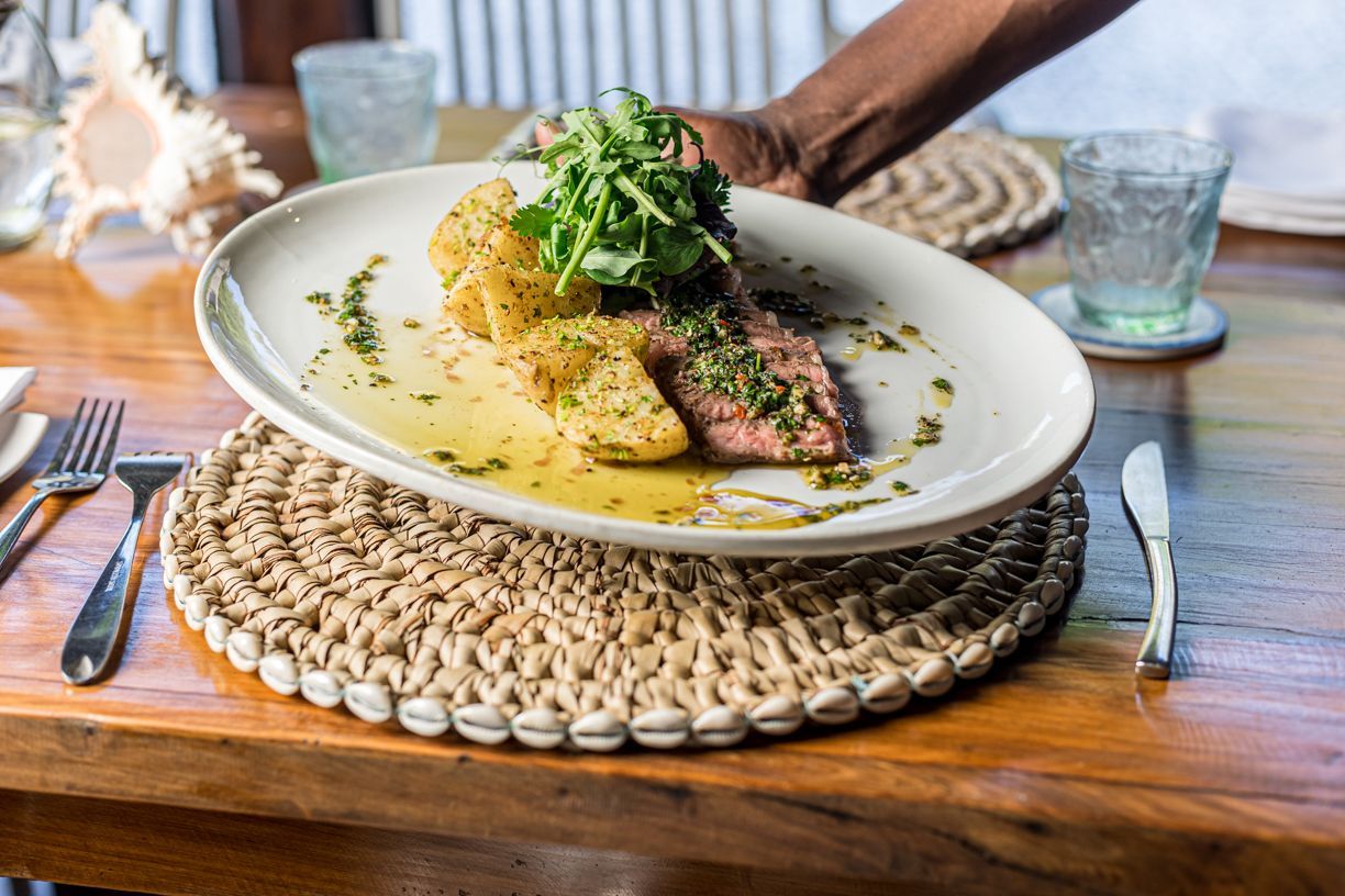 Hand placing a plated meal with greens and sauce on a woven placemat at a wooden table