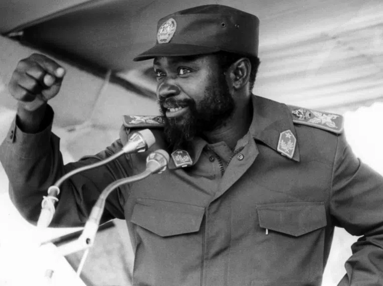 Samora Machel, in military uniform, speaks at a podium with his fist raised in the air.
