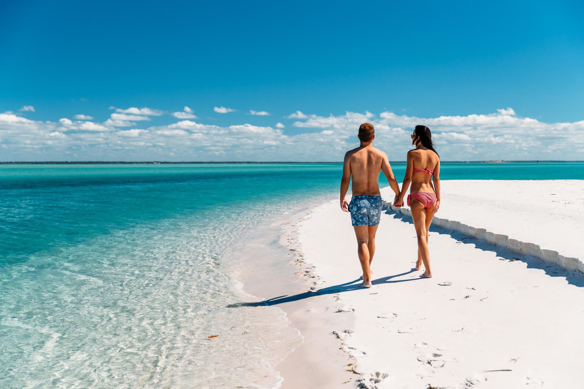 Two people walking hand in hand on a white sand beach beside turquoise water