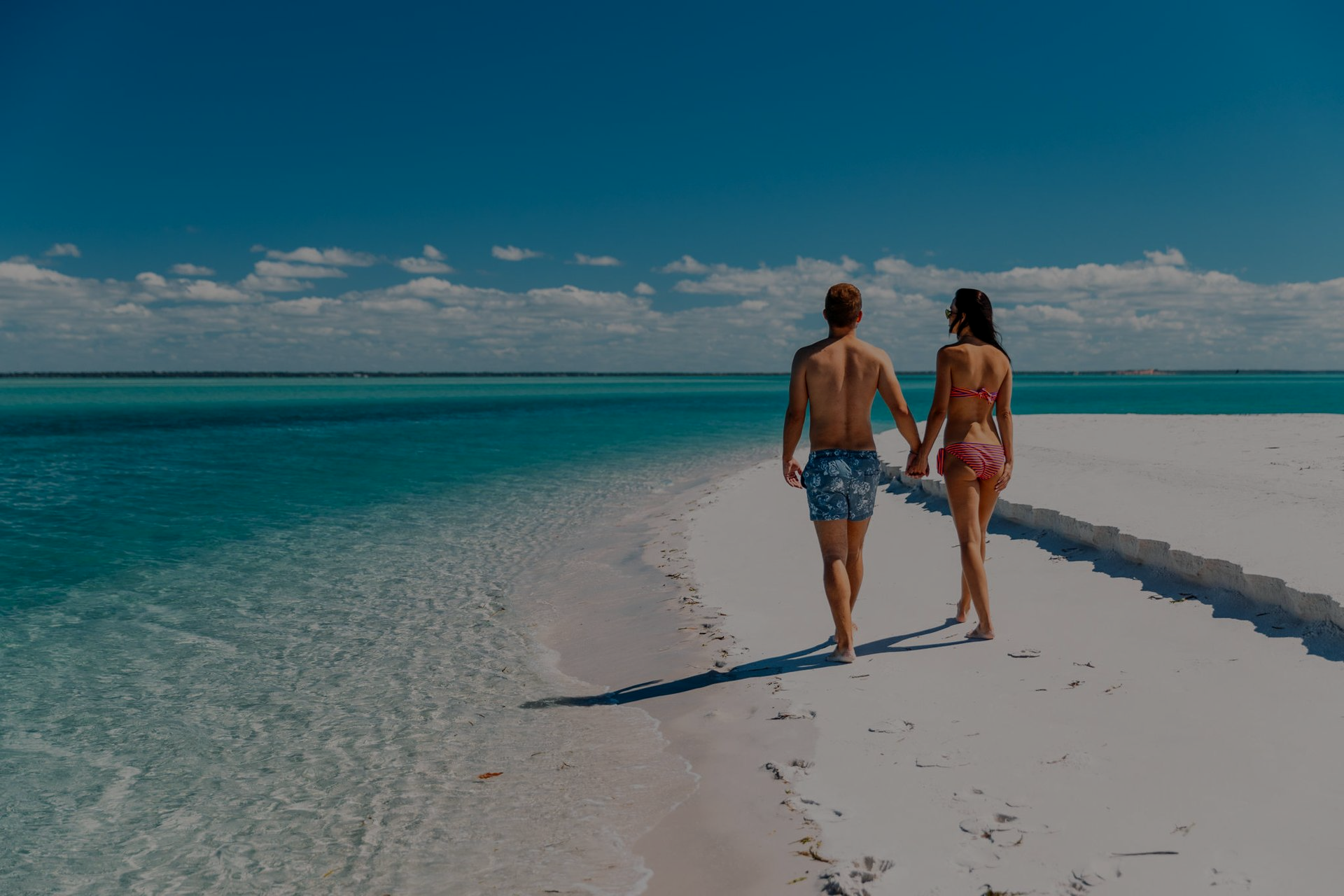Two people walking barefoot on a white sand beach beside turquoise water under a blue sky
