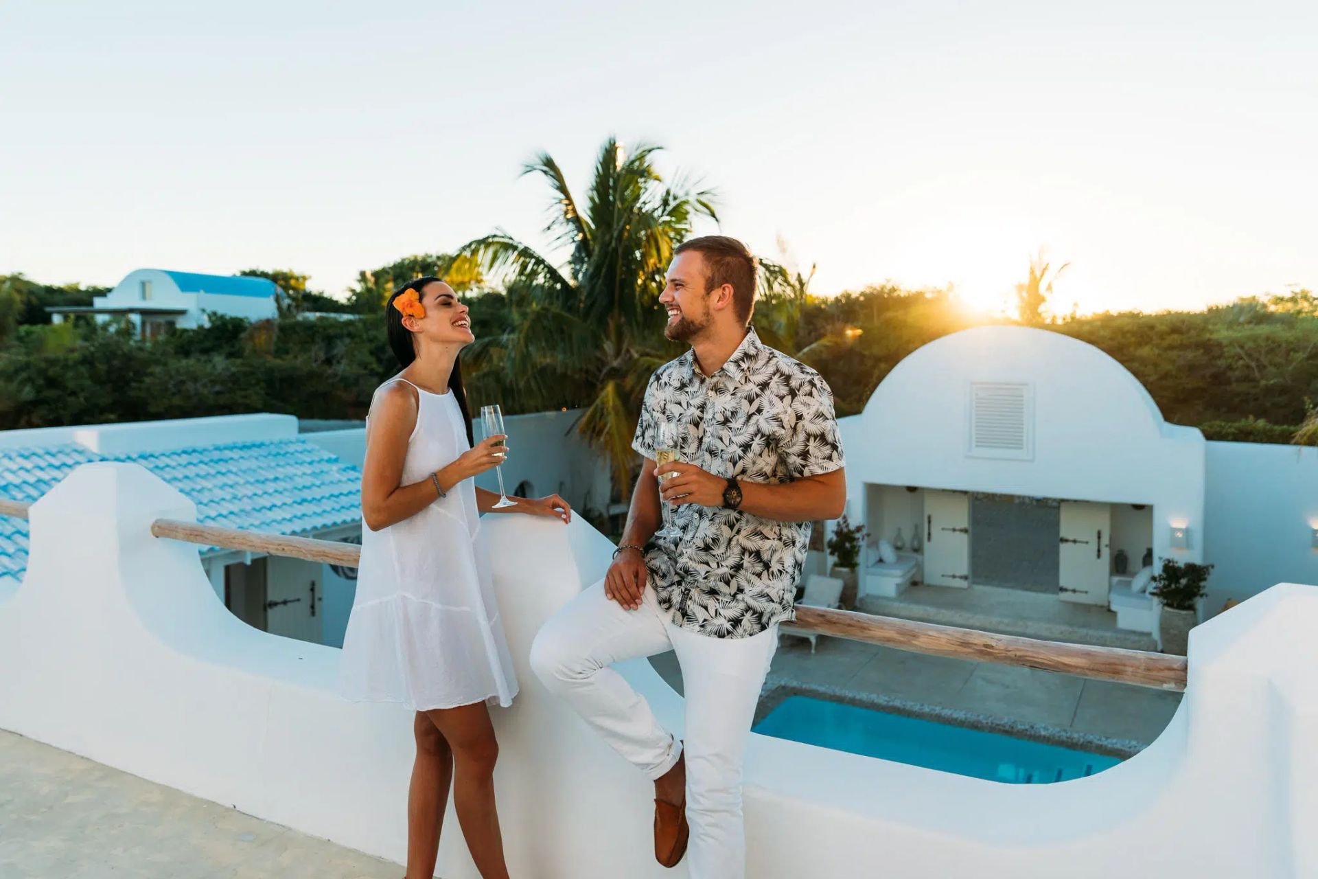 Two people in white outfits talk by a pool at a white resort with palm trees at sunset