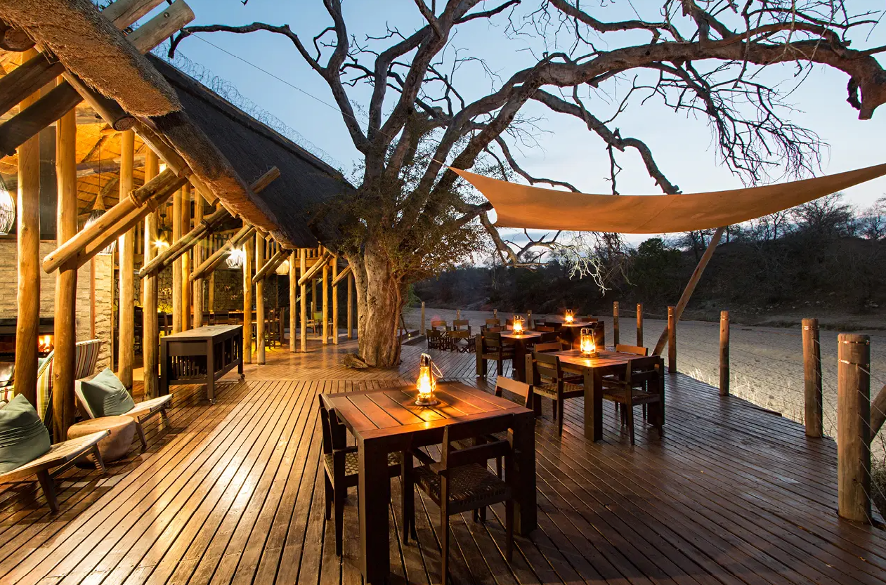 Wooden deck with candlelit tables beside a tree and river at dusk