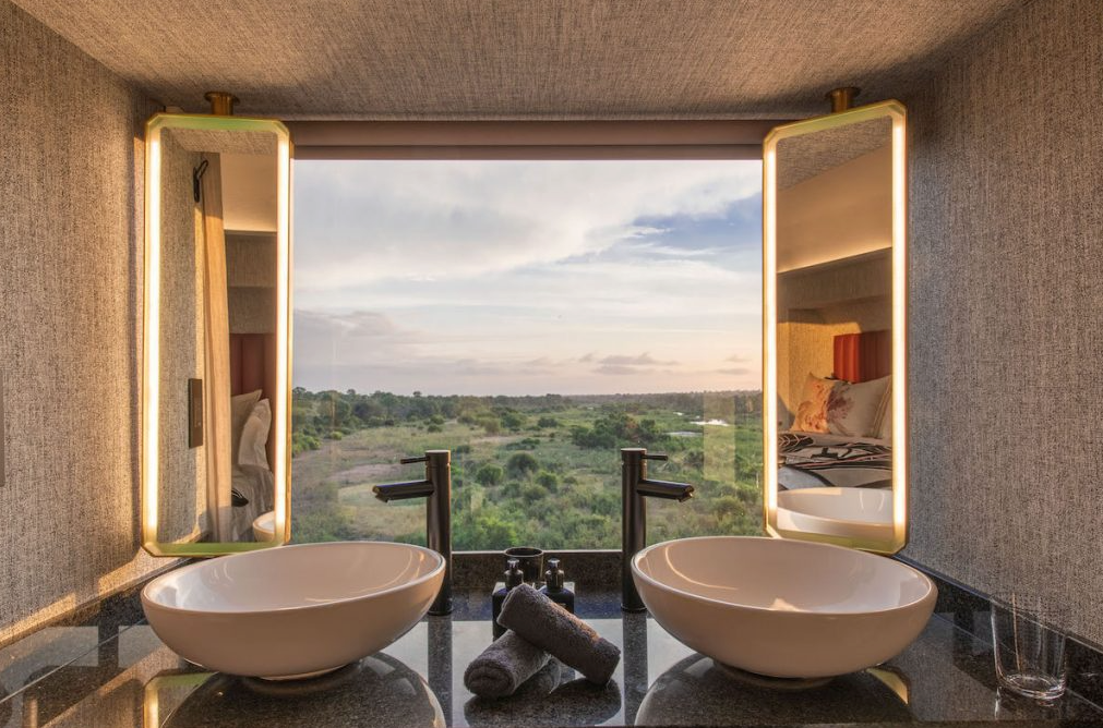 Spa bathroom with twin sinks and a large open window overlooking a green landscape at sunset