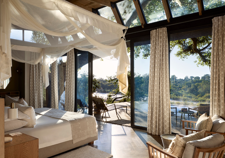 Luxury bedroom with sheer canopy bed, open glass walls, and river view at sunset