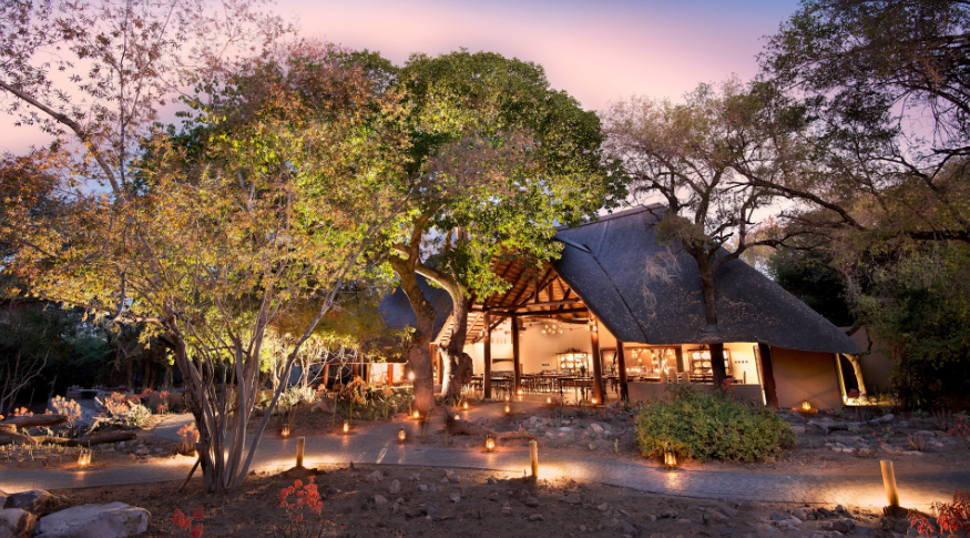 Thatched lodge among trees at dusk, lit warmly by firelight under a pink-purple sky.
