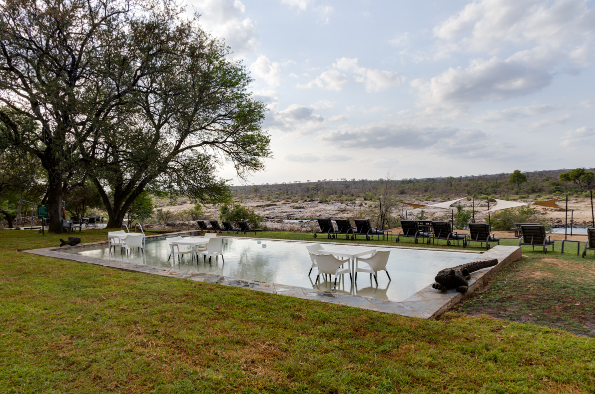 Outdoor patio with tables and chairs beside a reflective pool, overlooking a grassy landscape under cloudy skies