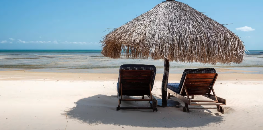 Two lounge chairs under a thatched umbrella on a sunny tropical beach by calm blue water.