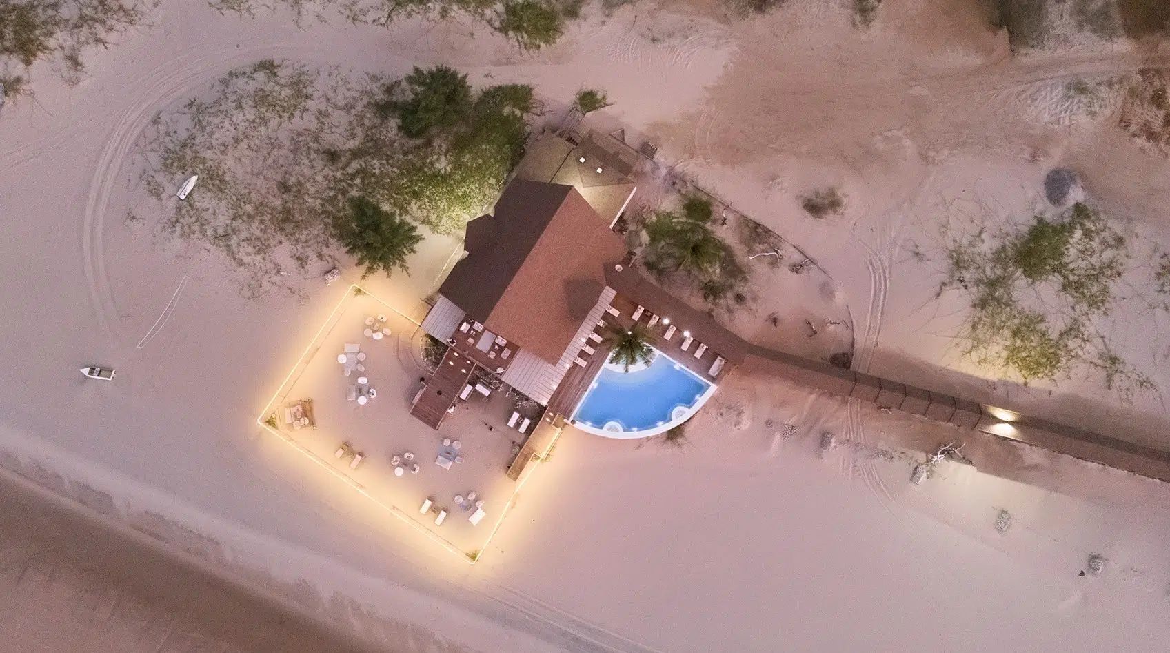 Aerial view of a lit beachside building with a blue pool, surrounded by sand and palm trees.