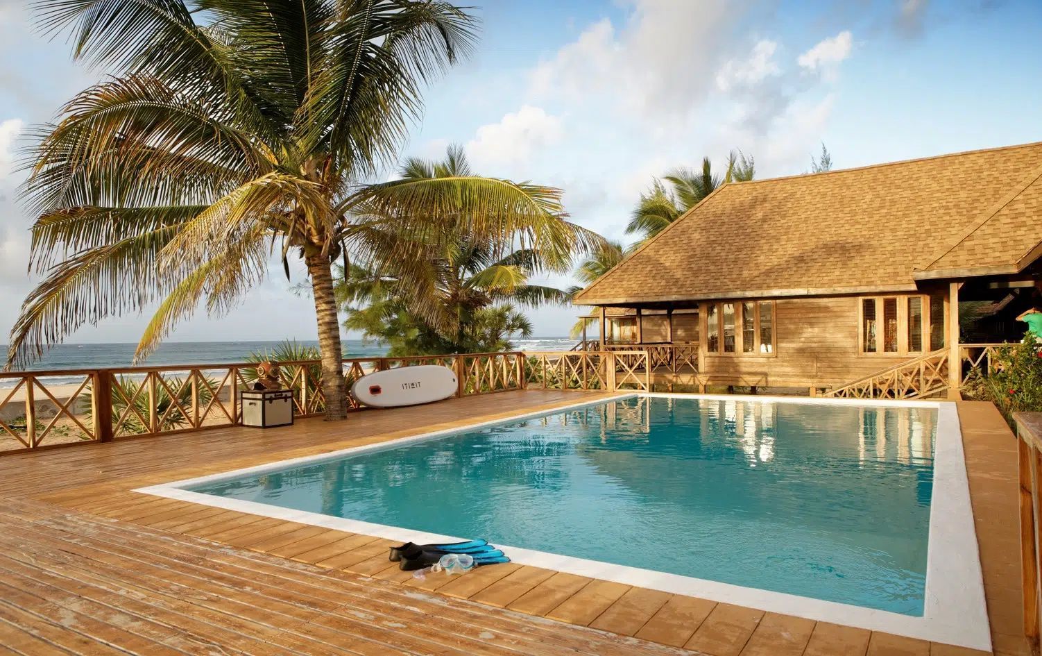 Resort pool beside palm trees and a thatched-roof lodge overlooking the ocean