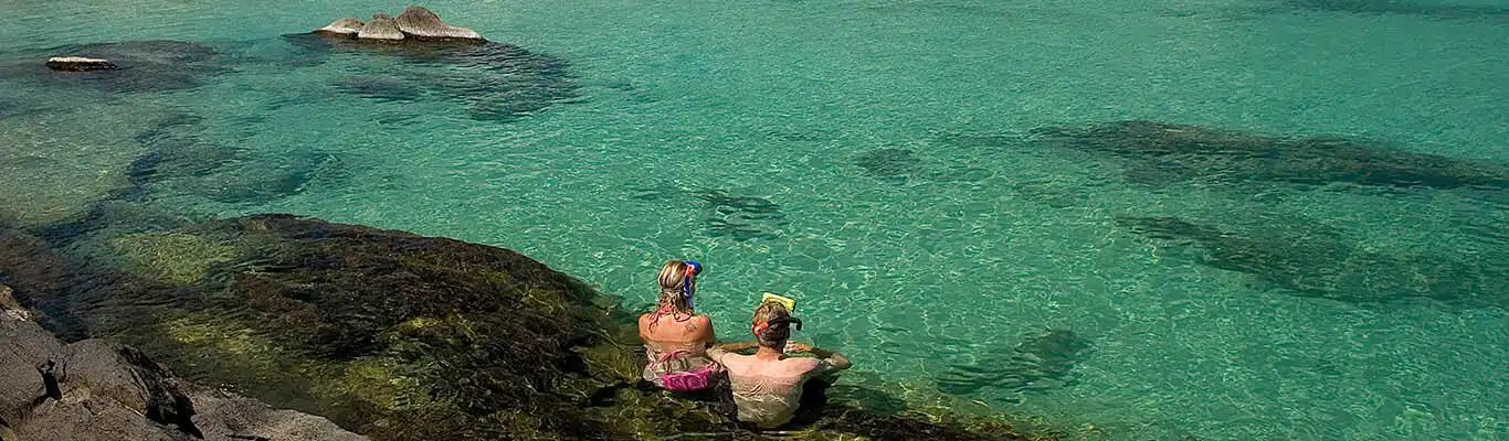 Two people sit on rocks beside clear turquoise water with a small boat in the distance.