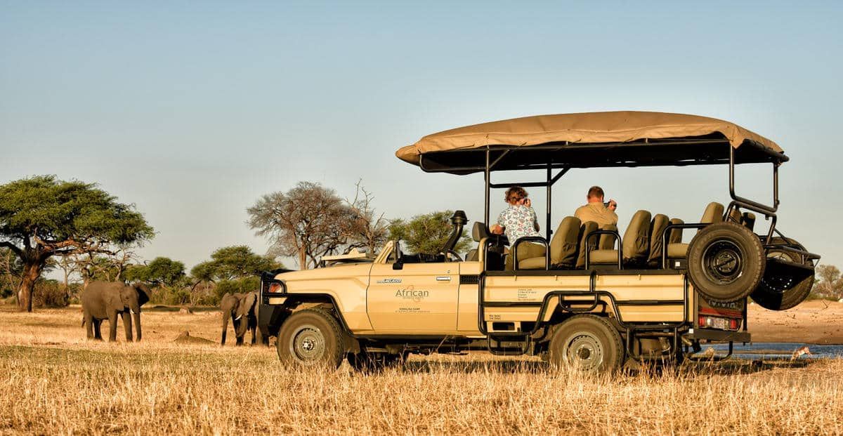 Safari vehicle with passengers in a dry grassland, elephants in the background at sunset